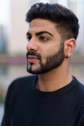 A young person with trendy men hairstyles Indian wearing a black ribbed sweater stands outdoors, with a blurred urban background of buildings and greenery.