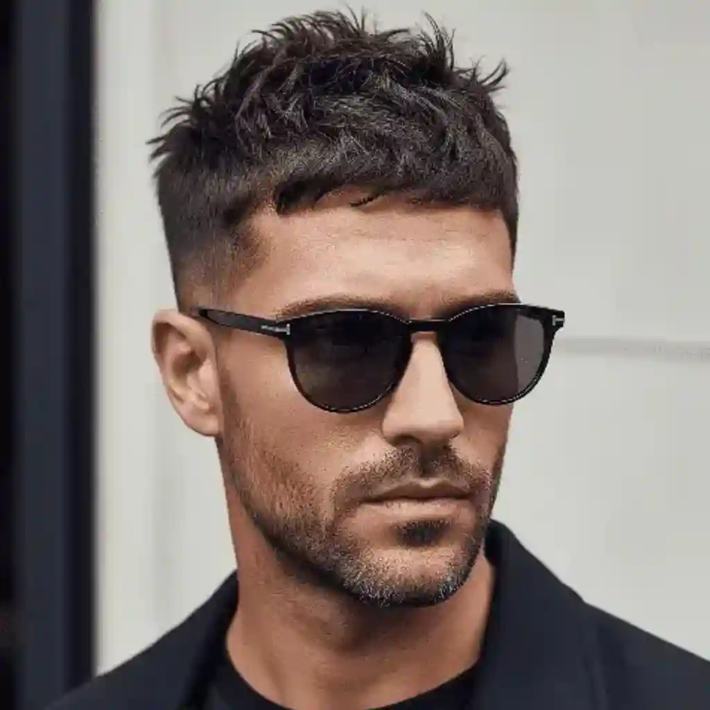 A young man with stylish, modern men short hairstyles featuring short, spiky hair, wearing dark sunglasses and a black jacket, posing against a light background.